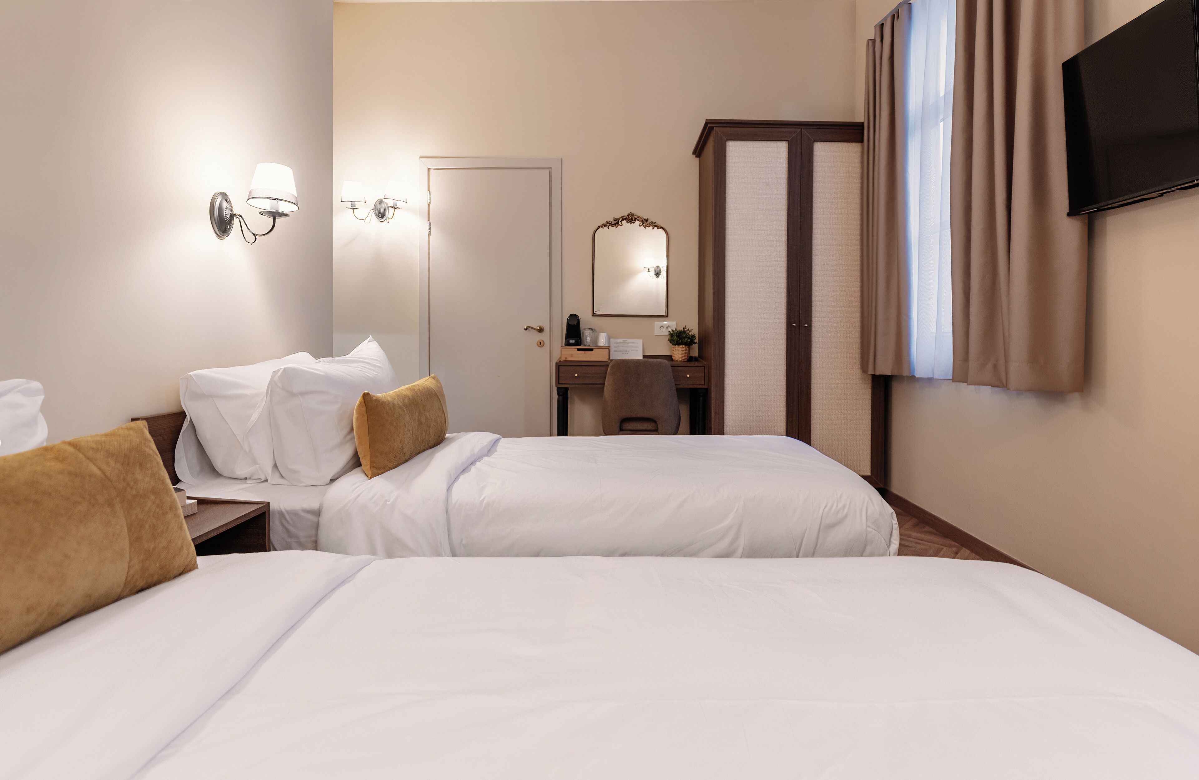 A hotel room with two beds, a desk with a chair, a mirror, a television, and a window with curtains.