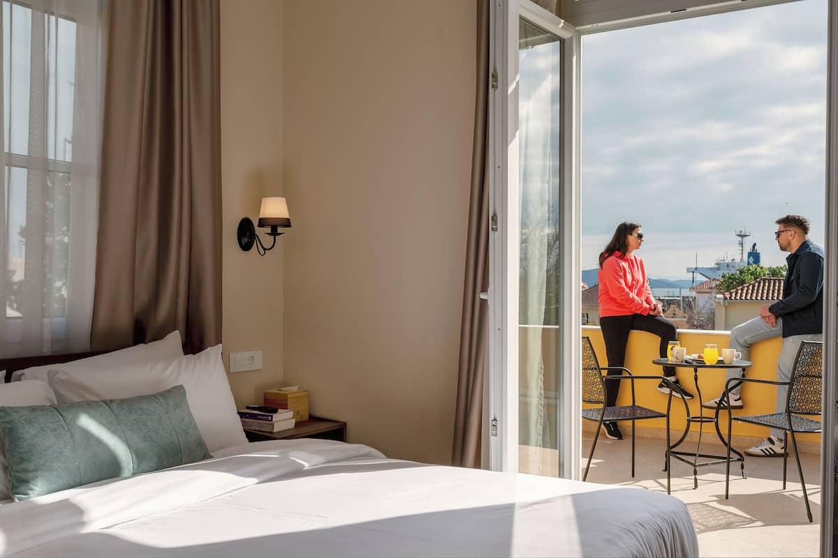A hotel room with a bed, a nightstand, a lamp, and a balcony with a table and chairs.