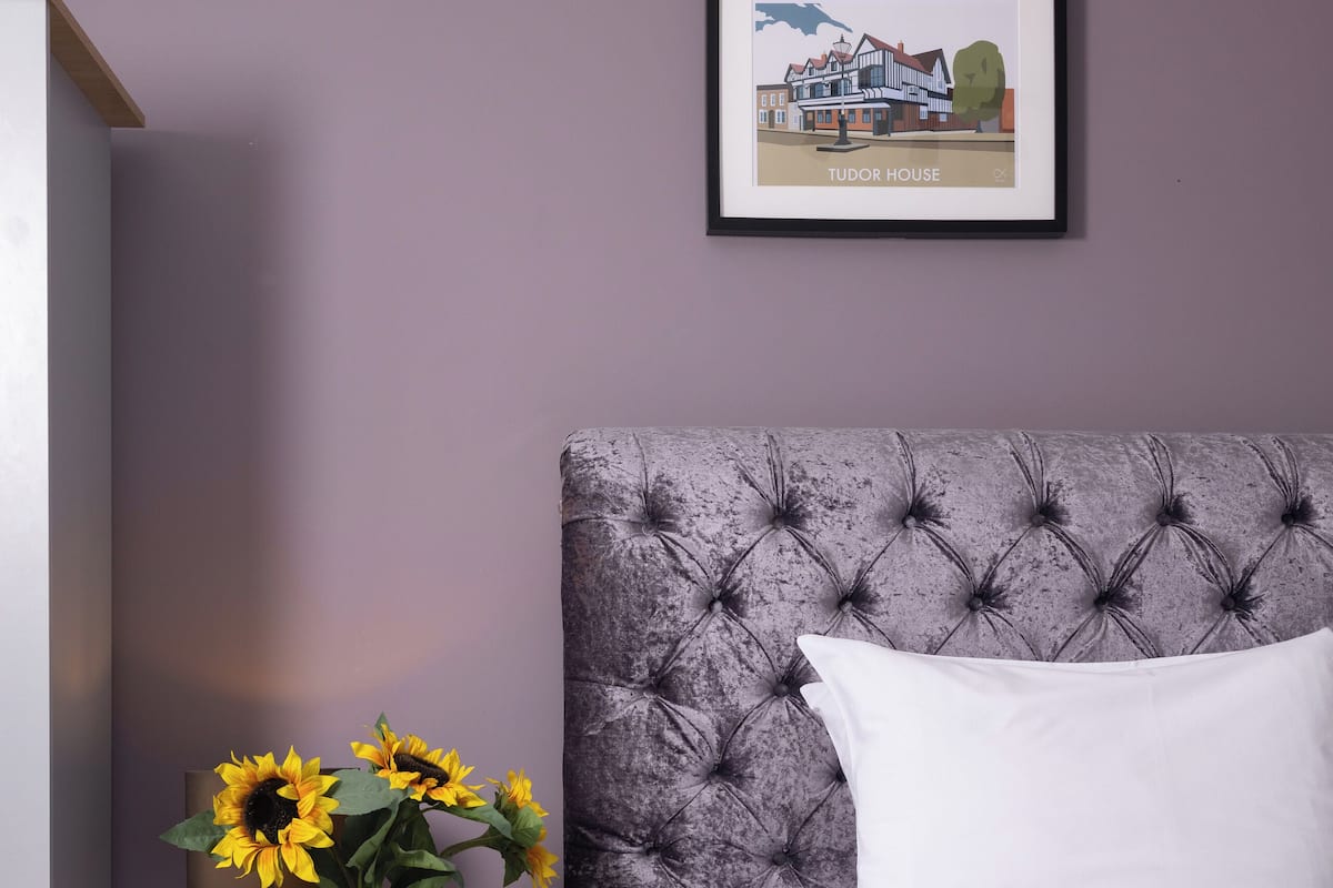 A bedroom with a tufted headboard, a white bed, a nightstand with a vase of sunflowers, and a framed picture of a building on the wall.