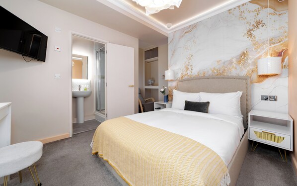 Una habitación de hotel moderna con una cama grande, una mesita de noche, un tocador, un televisor montado en la pared, un baño con lavamanos y ducha, y una pared decorativa con detalles dorados.