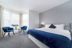 A hotel room with a large bed, two blue armchairs, a small table with a vase of flowers, and a nightstand with a lamp. - Harbour Hub Hotel (Southampton)