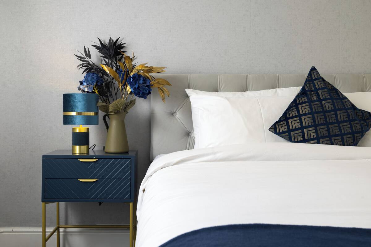 A neatly made bed with white linens and a patterned pillow, a bedside table with a lamp and a vase of flowers, and a wall with a subtle texture.