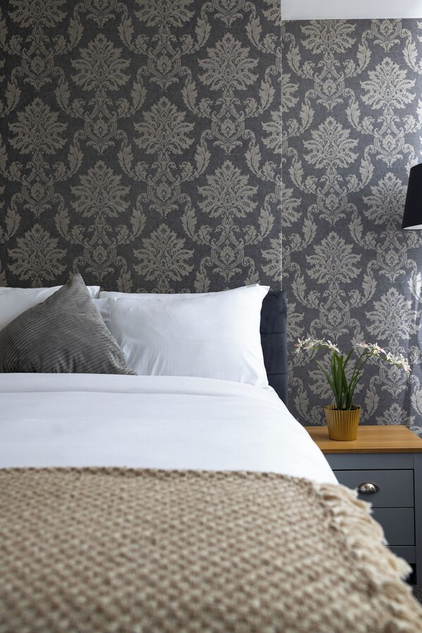 Una cama bien hecha con sábanas blancas y un almohadón gris, una mesita de noche con una planta en maceta y un papel tapiz oscuro con estampado.
