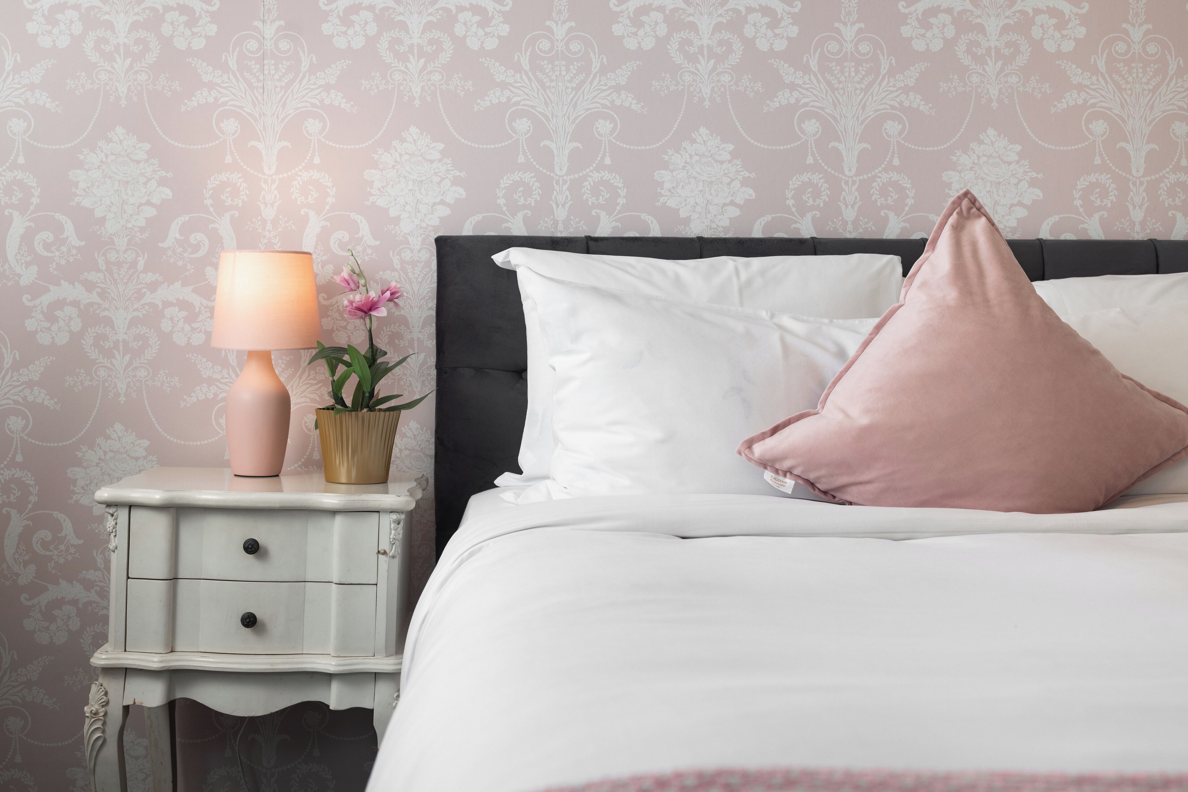 Una cama bien tendida con almohadas blancas y rosas, una lámpara de noche y una pequeña planta en maceta sobre una mesita blanca.