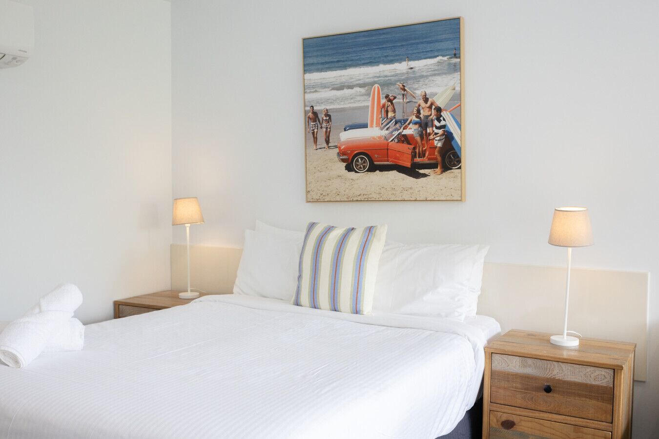 A neatly made bed with white linens, two bedside tables with lamps, and a beach scene painting on the wall.