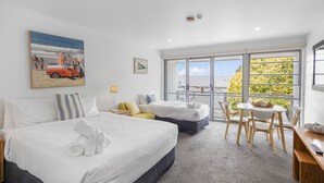 A hotel room with two beds, a balcony with a table and chairs, and a large window offering a view of greenery. - Bay View - The Trio (Lorne)