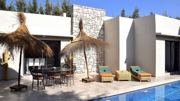 Un área de piscina exterior moderna con sillones reclinables, una mesa de comedor y un muro de piedra.