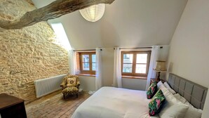 Una camera da letto con parete in pietra, un letto con biancheria bianca, due finestre con tende, una sedia e una lampada.