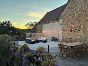 Un bâtiment en pierre avec un toit en pente, un espace extérieur aménagé avec un canapé et une table, et des guirlandes lumineuses.
