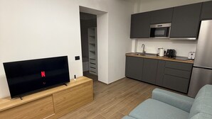 A modern living room with a flat-screen TV, a wooden entertainment unit, a grey sofa, and a kitchen area with dark cabinets and stainless steel appliances. - Adriano Space, a complete and functional apartment in the center of Modena (Modena)