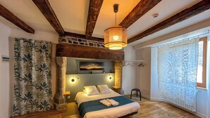 Un dormitorio con piso de madera, una cama con ropa blanca, una lámpara colgante y un ventanal con cortinas estampadas.