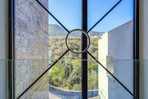 A view through a window with a geometric frame, revealing a stone wall and a landscape with trees and hills. - A luxurious specious 4 bedroom 3.5 bathrooms sea front modern villa (XIROPIGADO)