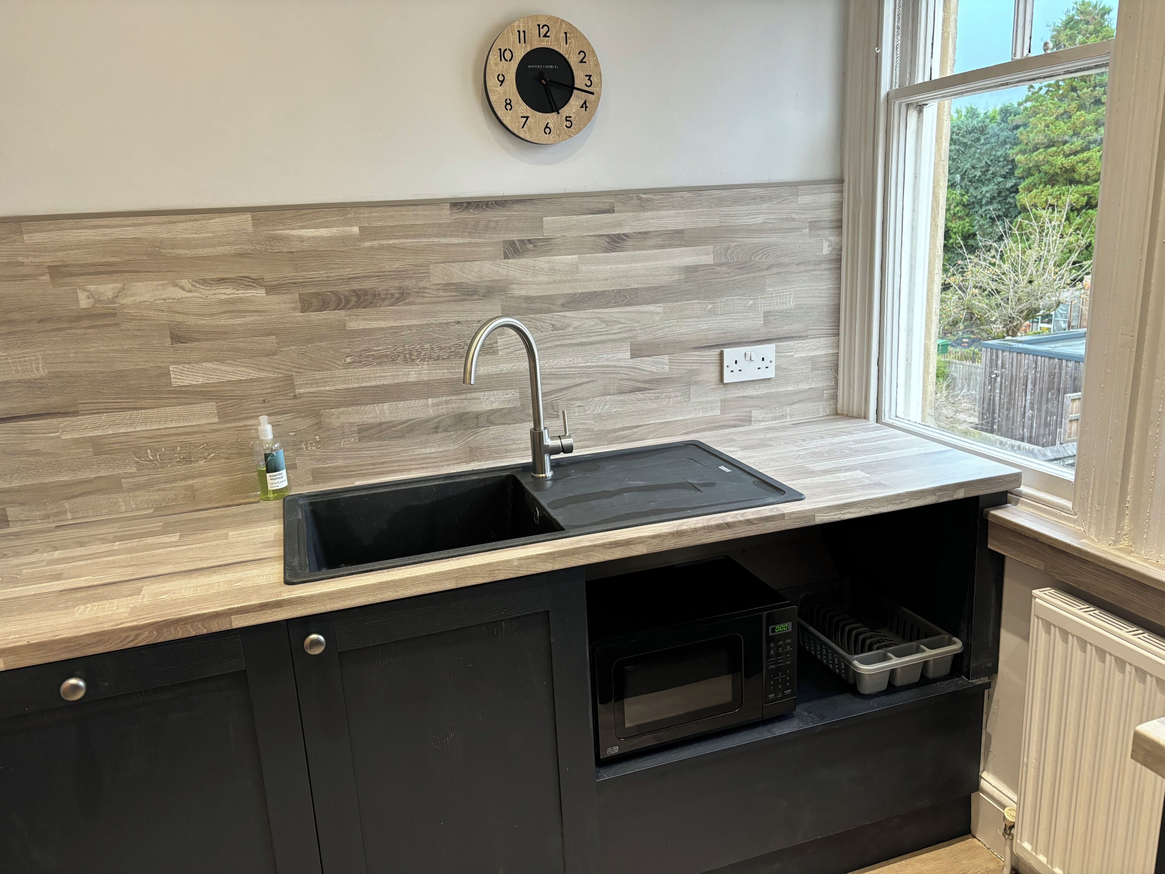 A kitchen with a wooden backsplash, a black sink, a stainless steel faucet, a microwave, and a window with a view of greenery outside.