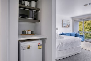 A modern hotel room with a bed, a blue sofa, a window with a view, a refrigerator, and a shelf with kitchen items. - Bay View - Green Vista (Lorne)