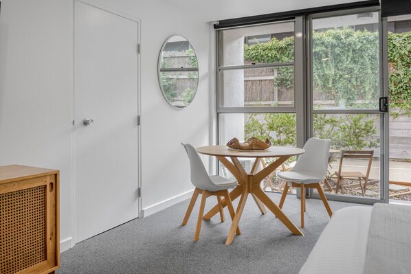A room with a large sliding glass door leading to a garden, a round table with chairs, and a wooden cabinet. - Bay View - Palm Springs (Lorne)