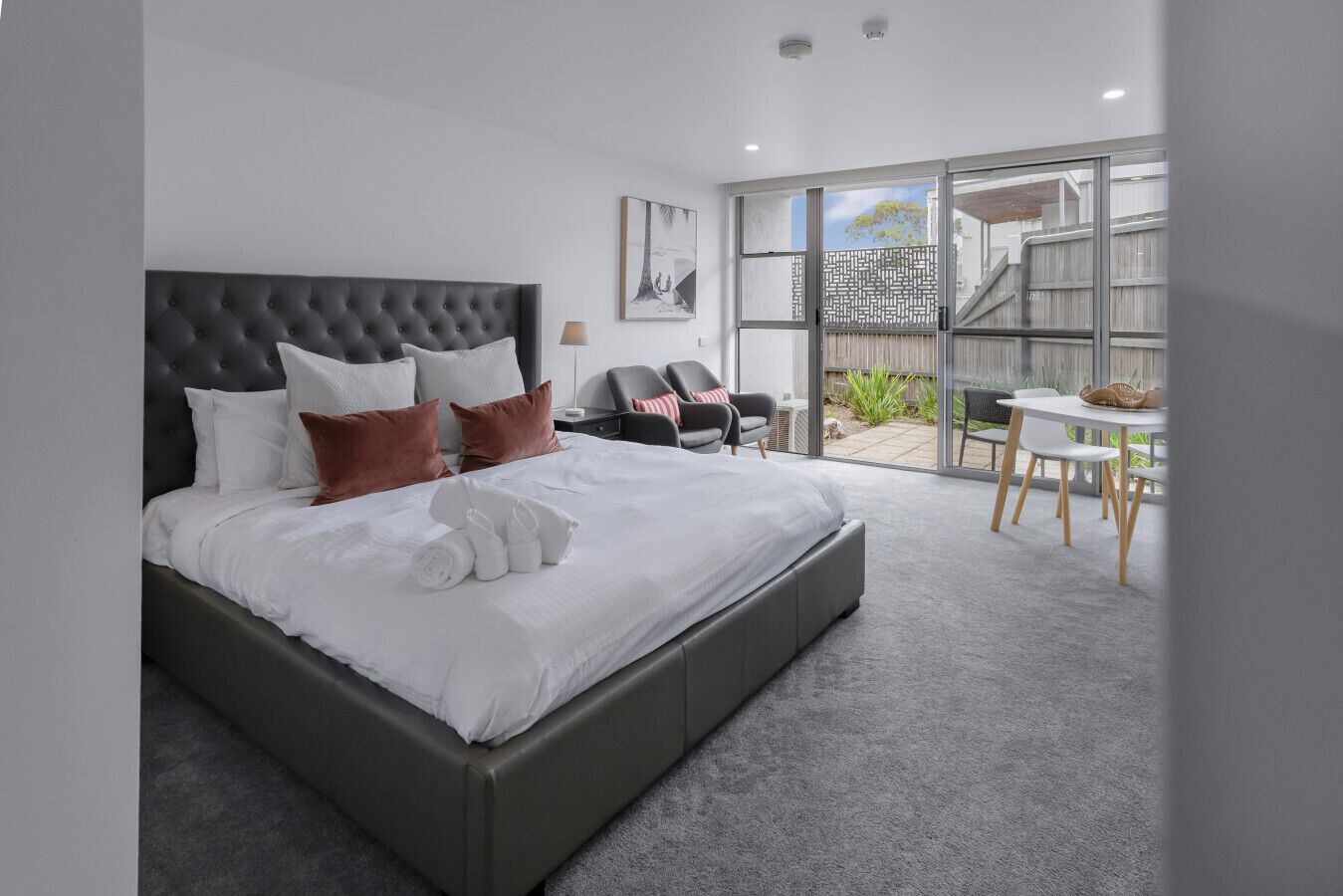 Une chambre moderne avec un grand lit, une chaise, une petite table et une porte-fenêtre coulissante donnant sur un espace extérieur.