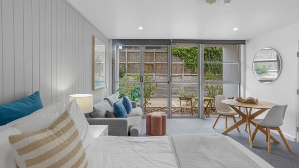 Un dormitorio moderno con una cama grande, un sofá, una mesa de comedor y vistas a un jardín a través de puertas de cristal.