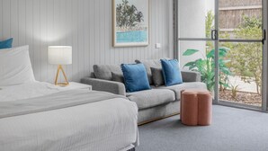 Un dormitorio con cama, sofá gris con cojines azules, un banquito redondo pequeño y un cuadro en la pared. Hay una ventana grande con vistas a la vegetación.