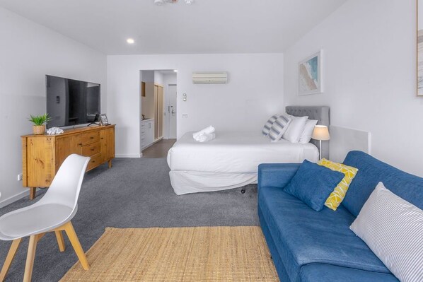 Une chambre d’hôtel moderne avec un lit, un canapé bleu, un téléviseur posé sur un meuble en bois et un petit bureau.