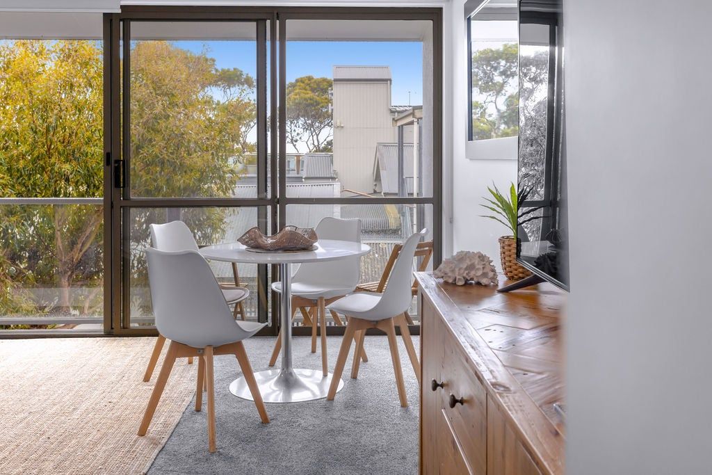 A dining area with a round table and chairs, a view through large glass doors, and a wooden cabinet with decorative items.