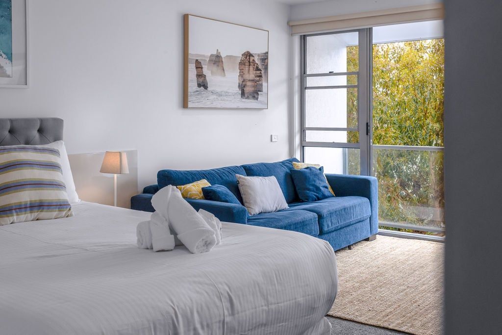 Een hotelkamer met een bed, een blauwe bank, een raam met uitzicht op bomen en een schilderij aan de muur.