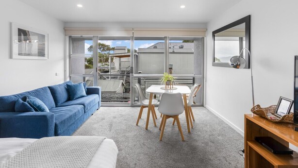 A modern living room with a blue sofa, a dining table with chairs, a small TV, and a balcony with a view of buildings and trees. - Bay View: Ocean Views (accessible) (Lorne)