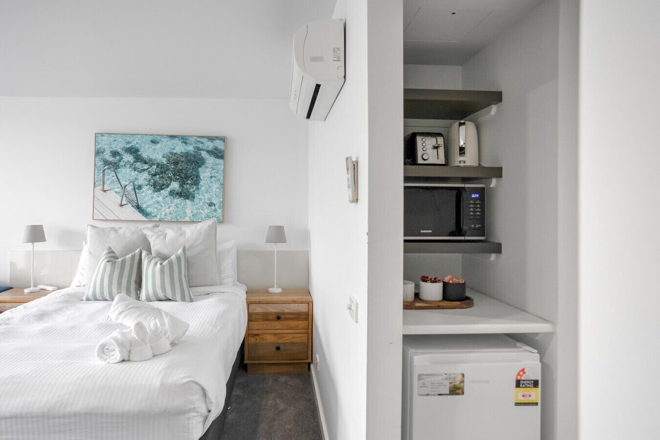 A modern hotel room with a neatly made bed, a small bedside table with a lamp, a wall-mounted air conditioner, a microwave, and a refrigerator.