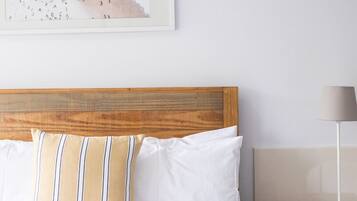 Ein ordentlich bezogenes Bett mit weißen Leinentextilien und gestreiften Kissen, ein hölzerner Kopfteil und ein Nachttisch aus Holz mit einer Lampe.