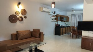 A modern living room with a sofa, a glass coffee table, and a TV. There is a kitchen area with cabinets and a dining table. - Villa Neesham Grande (Benaulim)