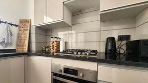 Une cuisine moderne avec des meubles blancs, une cuisinière en acier inoxydable et un étagère en bois où sont rangés les ustensiles de cuisine essentiels.
