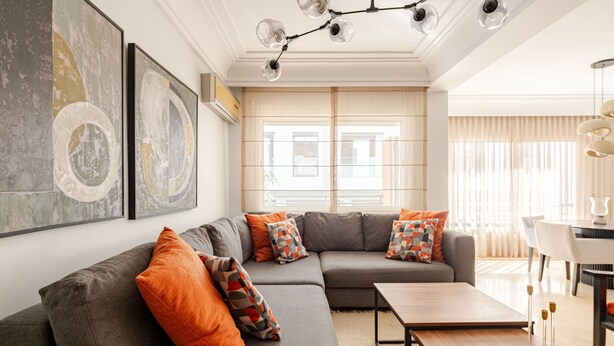 Ein modernes Wohnzimmer mit einem grauen Ecksofa, orangefarbenen Kissen, einem Holztisch und abstrakter Wandkunst.