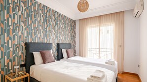 Ein Hotelzimmer mit zwei Betten, einem Wandteppich mit Muster, einem Fenster mit durchsichtigen Vorhängen, einem Nachttisch aus Holz mit einer Lampe und einer Deckenleuchte.