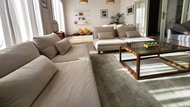 A modern living room with a beige sofa, a glass coffee table, and a large window with curtains. - The 31 Grand Theatre by IB Signature - Spacious Art Deco 1 BR Apt. (Casablanca)