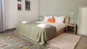 A neatly made bed with white and orange pillows, a green blanket, and folded towels on the floor. There are two framed posters on the wall, a bedside table with a lamp, and a patterned rug on the floor. - The 31 Grand Theatre by IB Signature - Spacious Art Deco 1 BR Apt. (Casablanca)