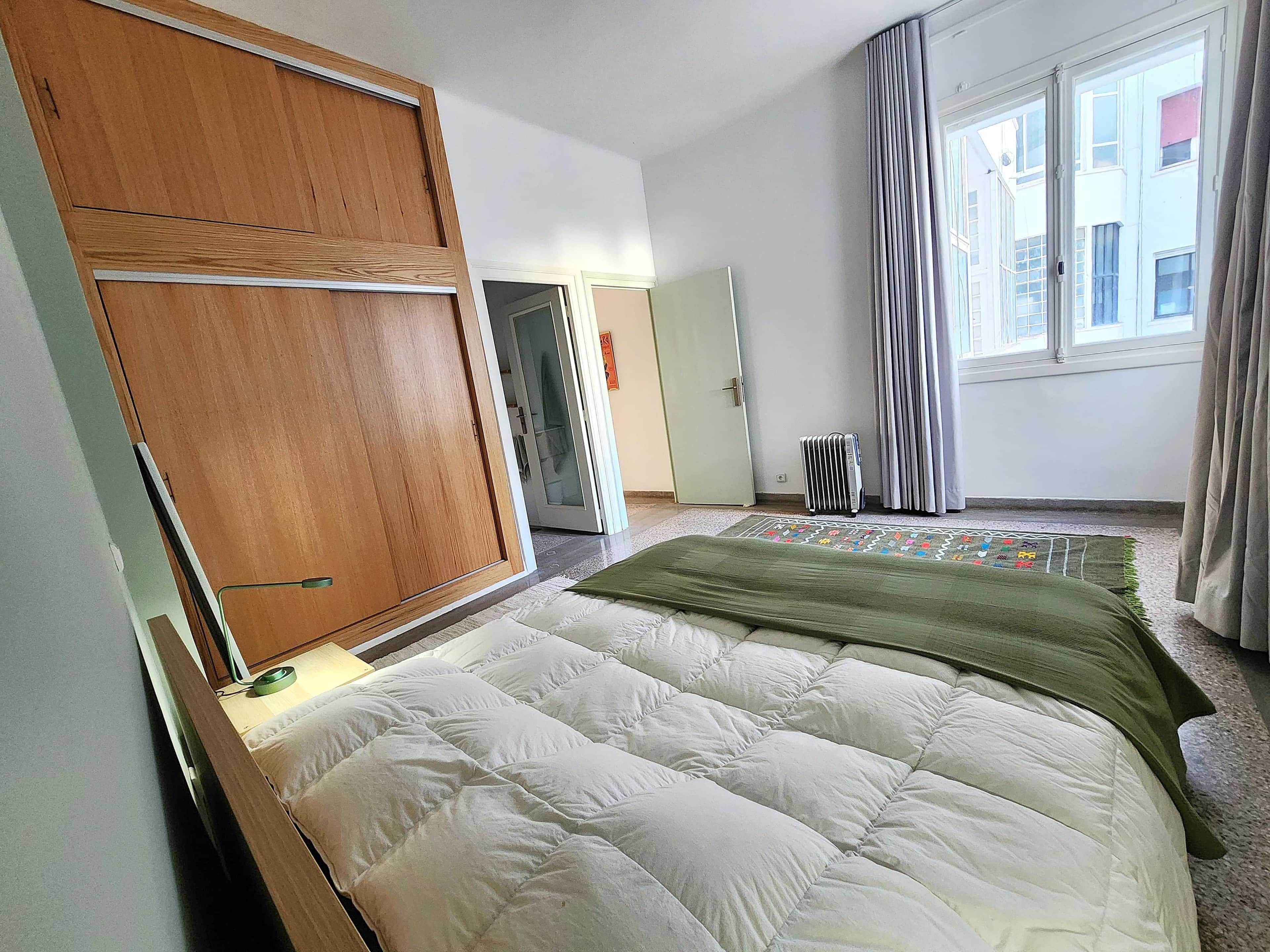 A bedroom with a bed, a wooden wardrobe, a radiator, and a window with curtains.