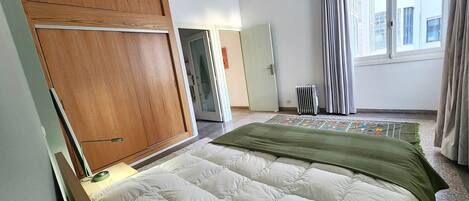 A bedroom with a bed, a wooden wardrobe, a radiator, and a window with curtains.