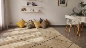 A living room with a beige carpet, a white dining table with chairs, a potted plant, and a bookshelf with books and decorative items. - The 31 Grand Theatre by IB Signature - Spacious Art Deco 1 BR Apt. (Casablanca)