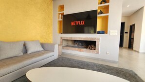 A modern living room with a grey sofa, a white coffee table, a fireplace, and a TV mounted on the wall displaying 'NETFLIX'. - Dar Bassidi by IB Signature - Luxury 5BR Villa -Heated Pool & Breakfast included (Marrakesh)