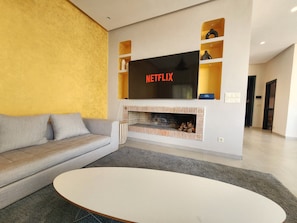 Ein modernes Wohnzimmer mit einem grauen Sofa, einem weißen Couchtisch, einem Kamin und einem an der Wand montierten Fernseher, der 'NETFLIX' anzeigt.