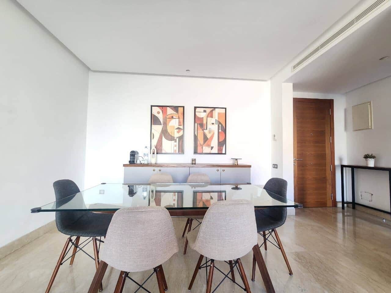Ein moderner Essbereich mit einem Glastisch, vier Stühlen, zwei abstrakten Bildern an der Wand und einer Holztür im Hintergrund.