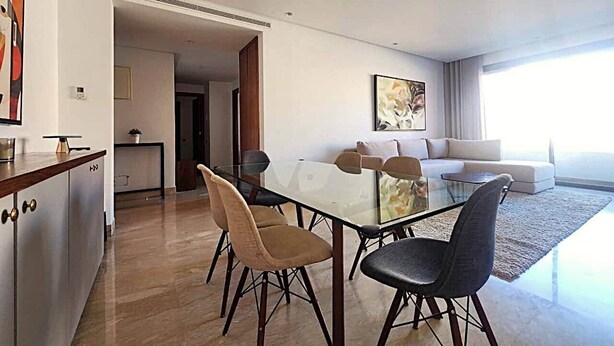 Un salon moderne avec une table à manger en verre, deux canapés et un coin cuisine doté de placards et d’un plan de travail.