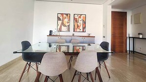 Un espace repas moderne avec une table en verre et quatre chaises, deux tableaux abstraits accrochés au mur, et une porte en bois à l’arrière-plan.