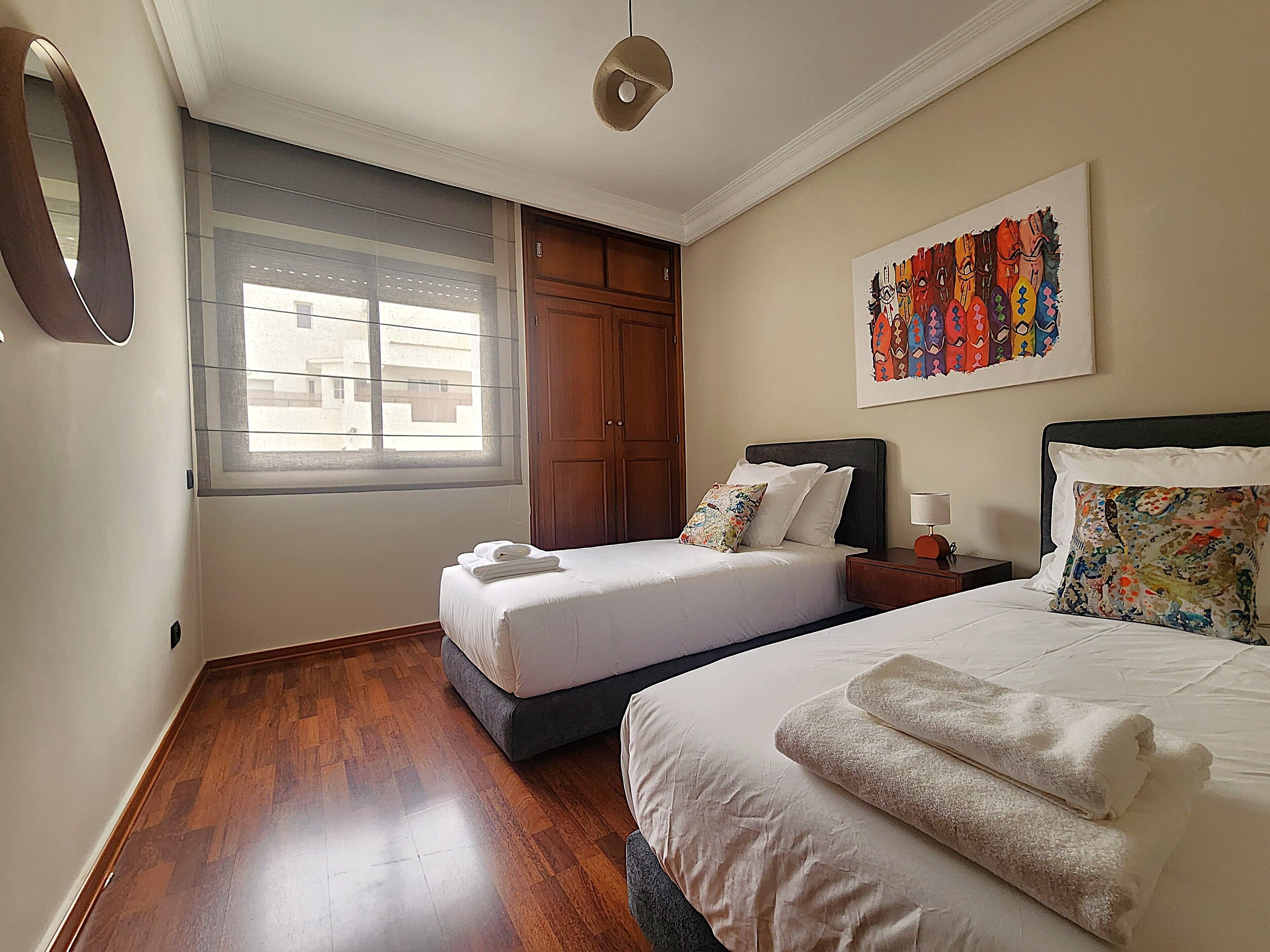 A hotel room with two beds, wooden flooring, a large window, and a colorful artwork on the wall.