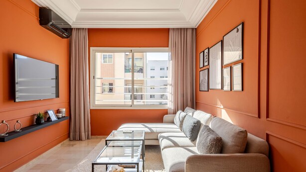 Ein Wohnzimmer mit orangefarbenen Wänden, einem Flachbildfernseher, einem Sofa, einem Couchtisch und einem großen Fenster mit Vorhängen.