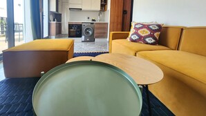 Ein modernes Wohnzimmer mit Sofa, Couchtisch und Essbereich. Im Hintergrund ist eine Küche mit einer Waschmaschine zu sehen.