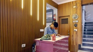 A reception desk with a person writing, wooden paneling on the walls, a ceiling fan, and a staircase in the background. - Basil Inn near To Taj Mahal (Agra)
