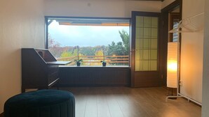 Una habitación con un ventanal grande, una computadora portátil sobre un escritorio, un taburete azul oscuro y una lámpara de pie.