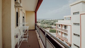 Un balcón con barandilla de metal, una silla blanca y una mesa. Se ve una vista de la ciudad con edificios y árboles.