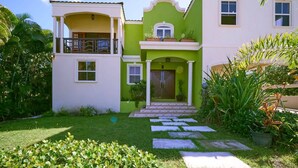 Una casa de dos pisos con fachada verde y blanca, un balcón en el segundo piso y un jardín bien cuidado con piedras que guían el camino hacia la entrada.