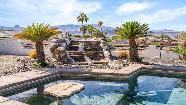 A swimming pool with a rock waterfall feature, surrounded by palm trees and a desert landscape in the background. - The Sun Chaser, 4 bdr. Pool, Spa and Endless Views (Lake Havasu City)